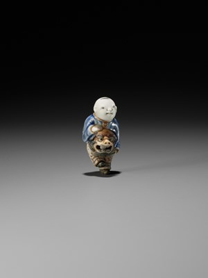 Lot 301 - A HIRADO GLAZED PORCELAIN NETSUKE OF A BOY WITH SHISHIMAI MASK