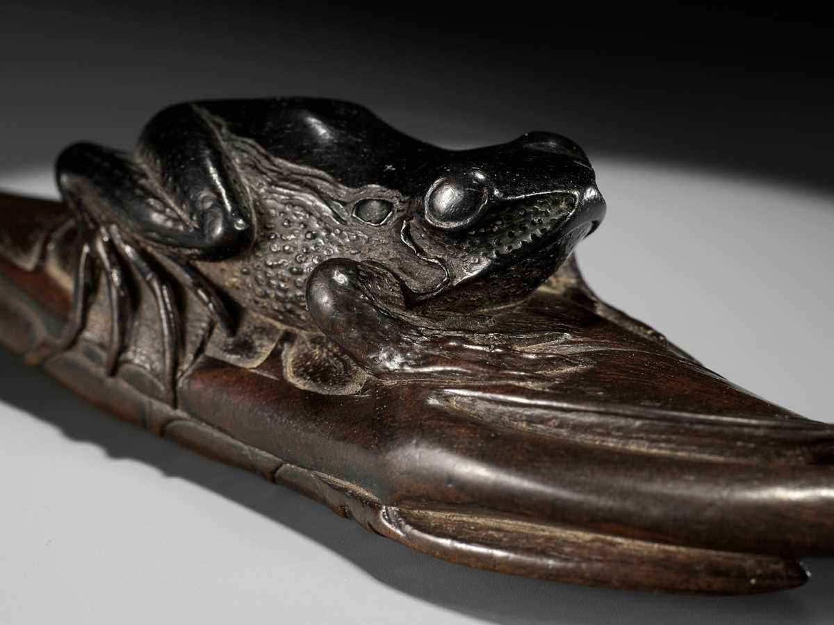 Lot 162 - KANMAN: AN EXCEPTIONAL AND LARGE KUROGAKI (BLACK PERSIMMON) WOOD NETSUKE OF A FROG ON A LOTUS LEAF
