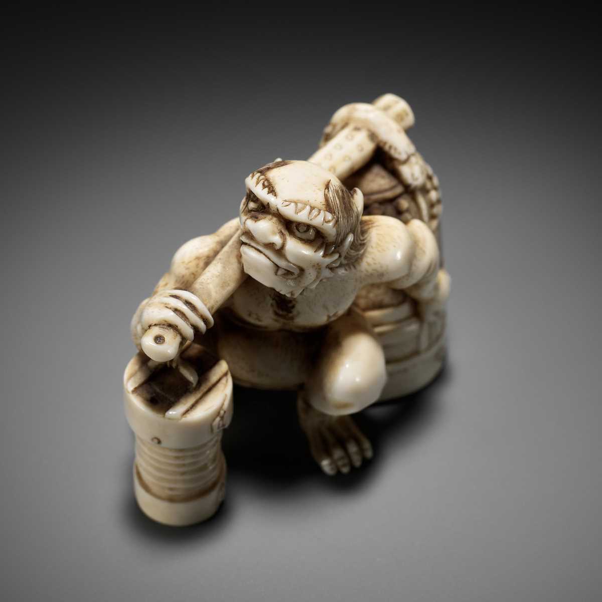 Lot 500 - RYOZAN: AN AMUSING IVORY NETSUKE OF AN ONI CARRYING A BONSHO AND CHOCHIN, CHOCHIN NI TSURIGANE