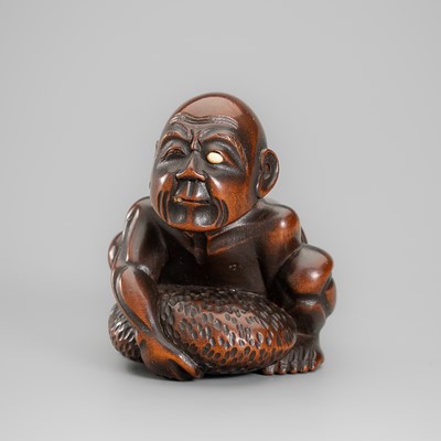 GYOKKEI: A FINE INLAID WOOD NETSUKE DEPICTING A BLIND MASSEUR LIFTING A CHIKARAISHI